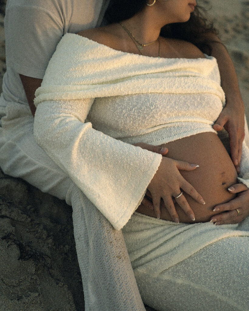 Photo of man and woman sitting on beach together, holding pregnant belly