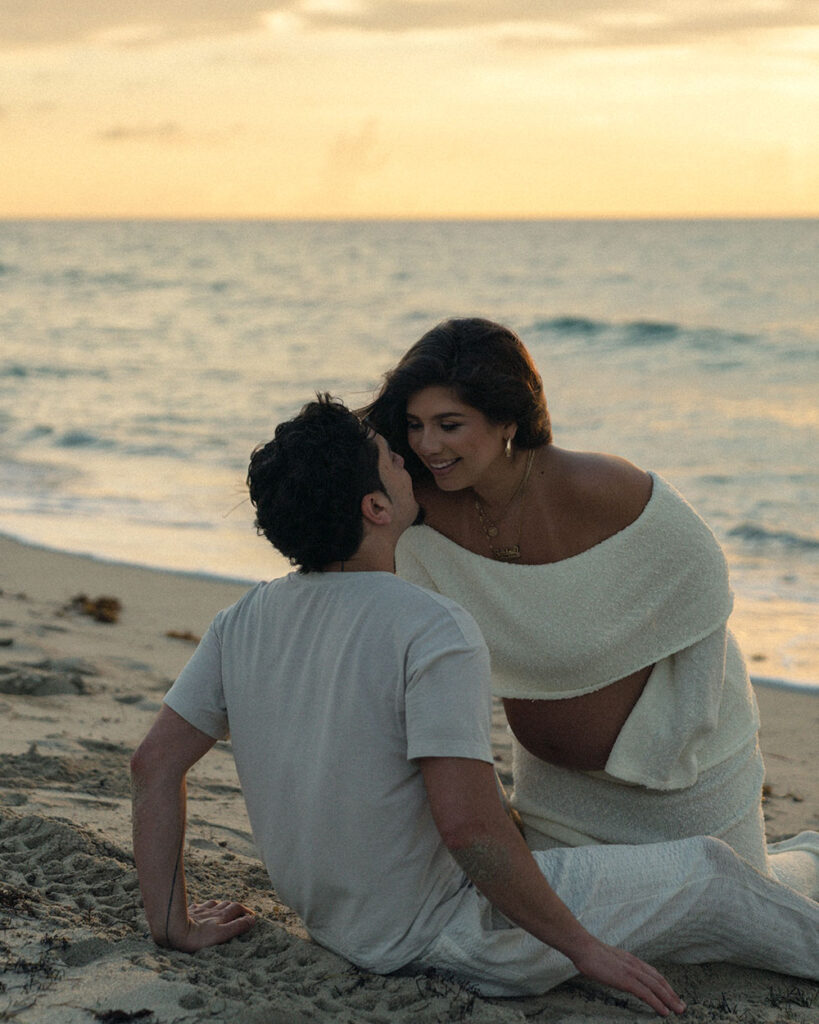 Pregnant woman leaning in to kiss man who is sitting on the shore of the beach